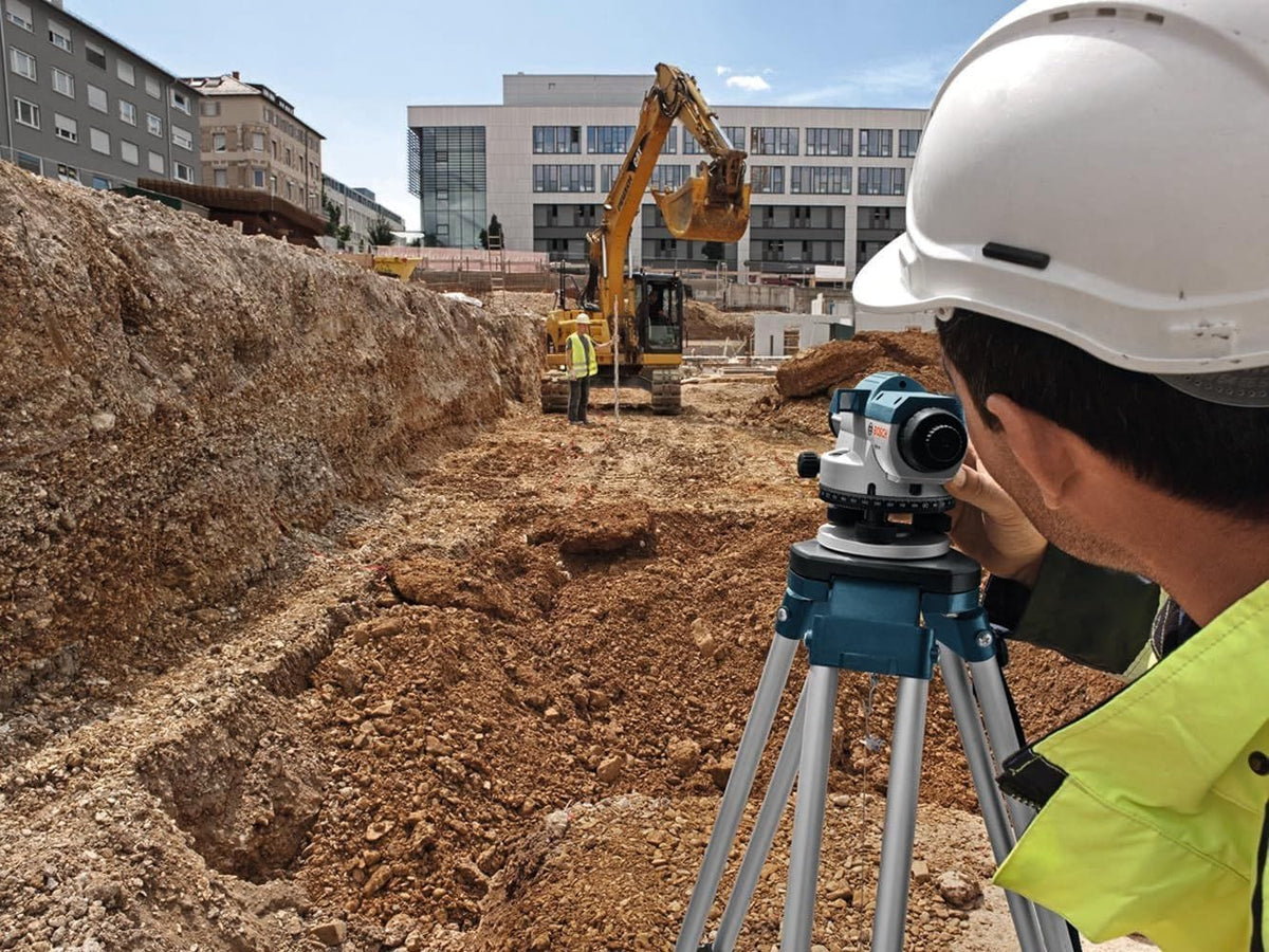 An outdoor, real-world application shot showing a construction site surveyor wearing a white hard hat and high-visibility vest as he looks through a tripod-mounted Bosch GOL 26 automatic optical level tool, adjusting the alignment focus while another worker holds a leveling rod in the background near a yellow excavator at a foundation grading site.