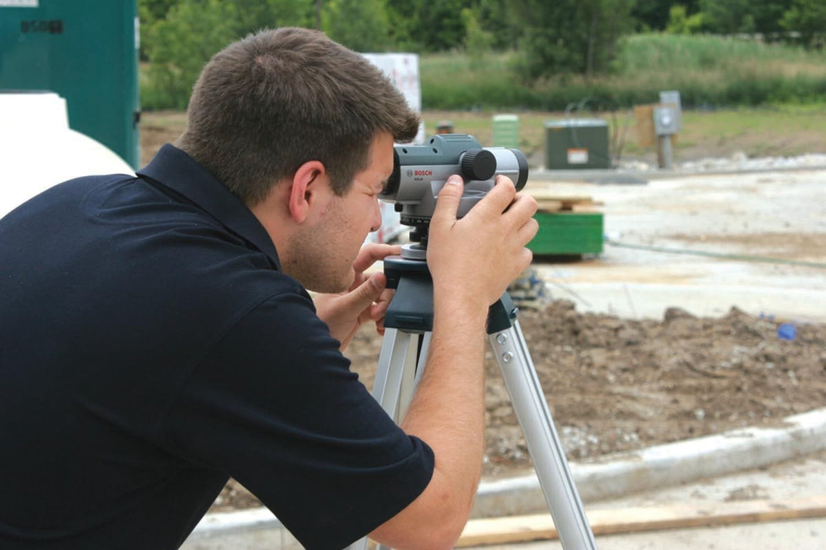 A side-profile action shot of a professional measuring site layouts using the Bosch GOL26 26x automatic optical level kit, with the instrument secured tightly to an aluminum construction tripod on an outdoor land development project.