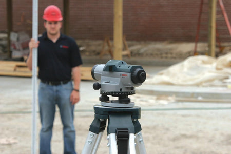 The Bosch GOL26 automatic optical level site surveyor measuring tool positioned prominently in the foreground on an aluminum tripod, with a construction worker holding a vertical telescoping leveling grade rod standing out of focus in the background of a commercial building project.