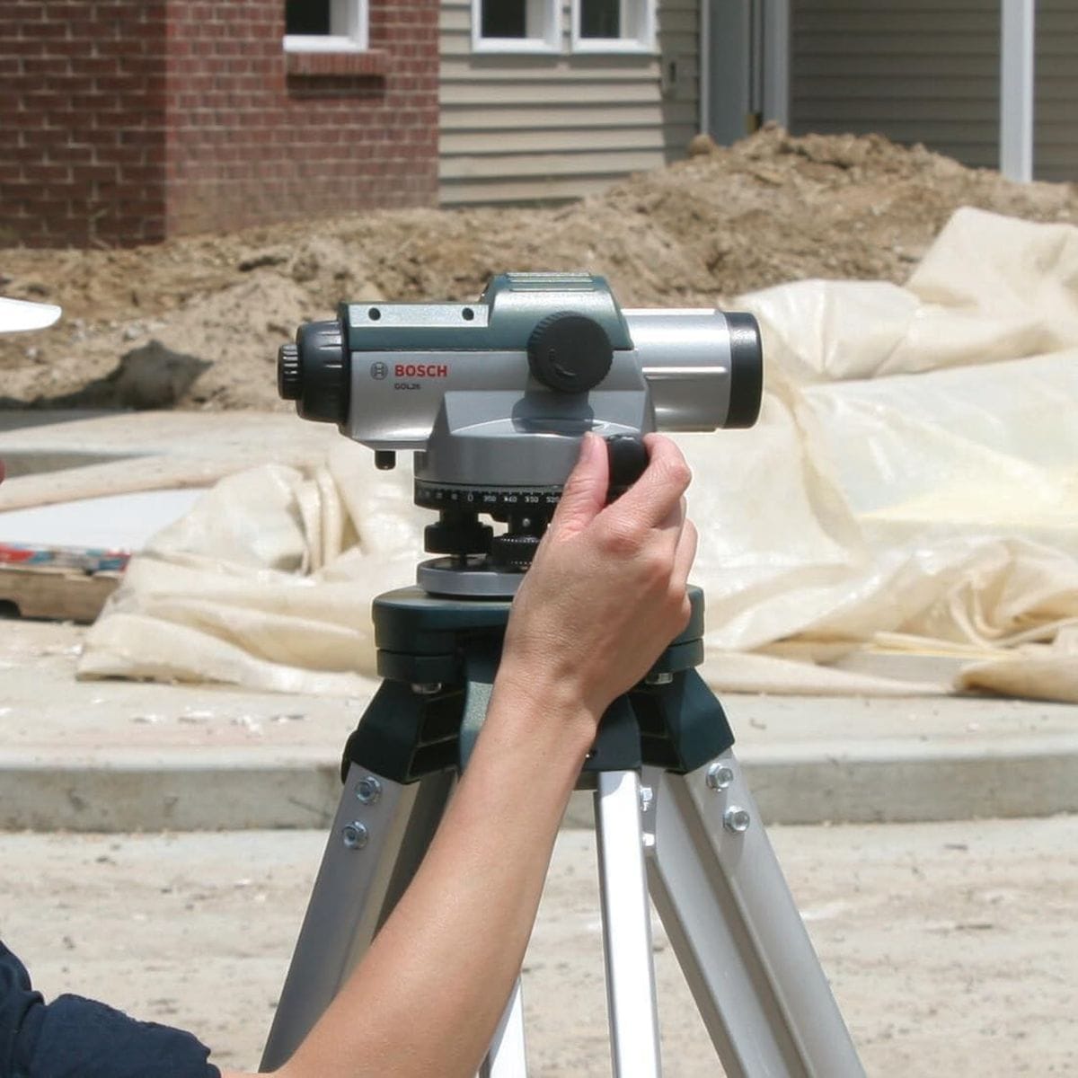 A close-up shot of a tradesperson's hand adjusting the horizontal drive screw on the Bosch GOL26 automatic optical level surveyor alignment tool to fine-tune the lens angle positioning during an outdoor residential concrete pour leveling setup.