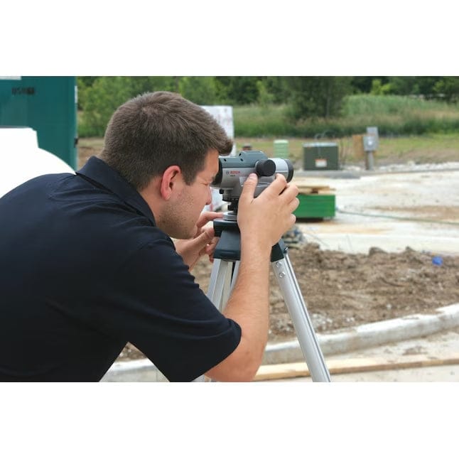 Person using an optical level mounted on a tripod at a construction site; measuring elevation with surrounding infrastructure and materials.