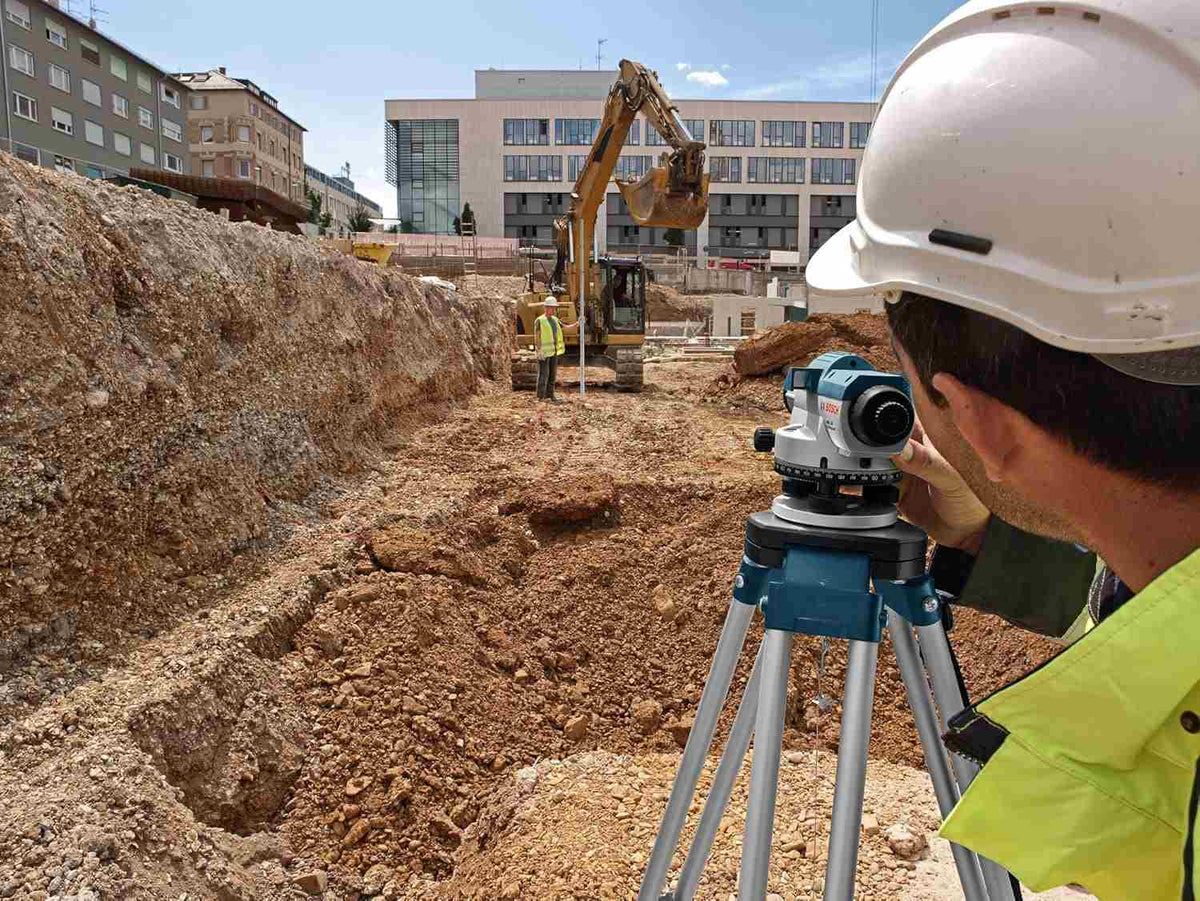 Worker using surveying instrument on tripod near excavator at urban construction site; buildings visible in background.