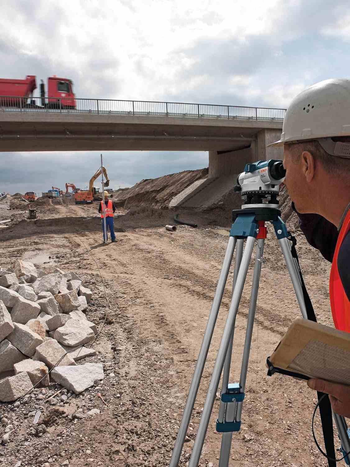 Surveyor operating tripod-mounted instrument at construction site; bridge, excavators, and second worker visible in background.