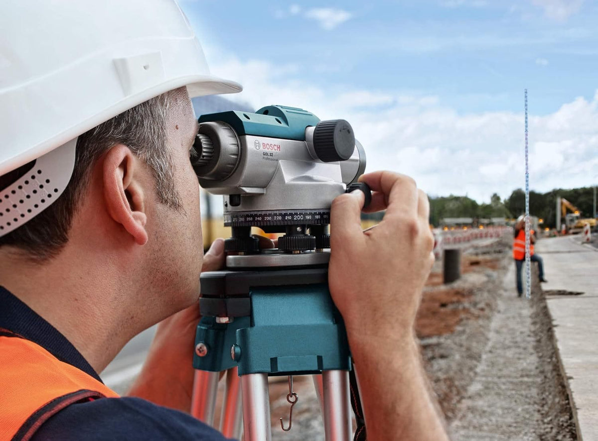 Construction worker using Bosch optical level on tripod to measure elevation; another worker stands near measuring rod.