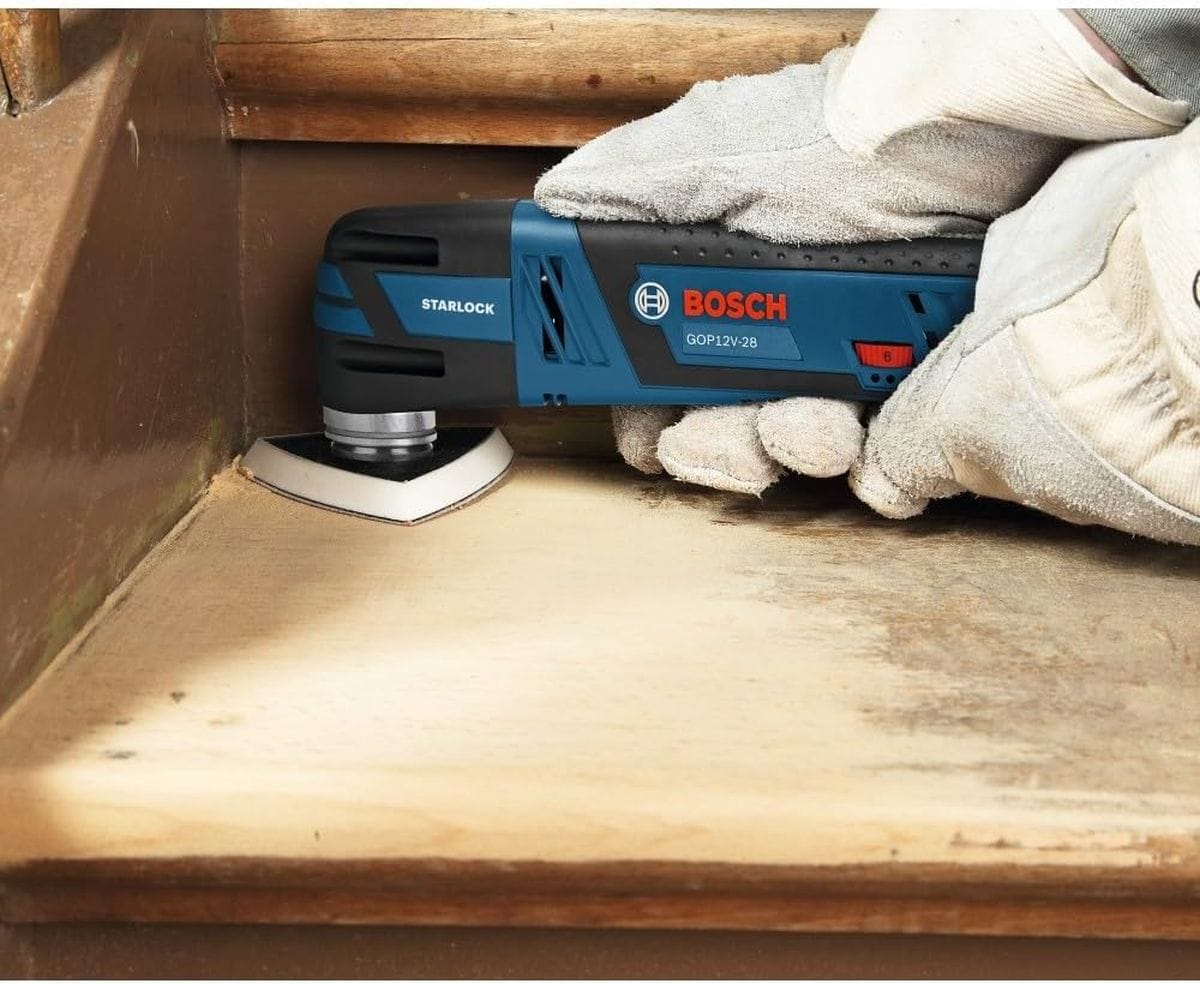 An active jobsite view of a worker wearing heavy-duty safety gloves using the Bosch 12V Max oscillating multi-tool equipped with a delta triangle sanding pad accessory to smoothly finish a tight internal corner on a wooden staircase step.
