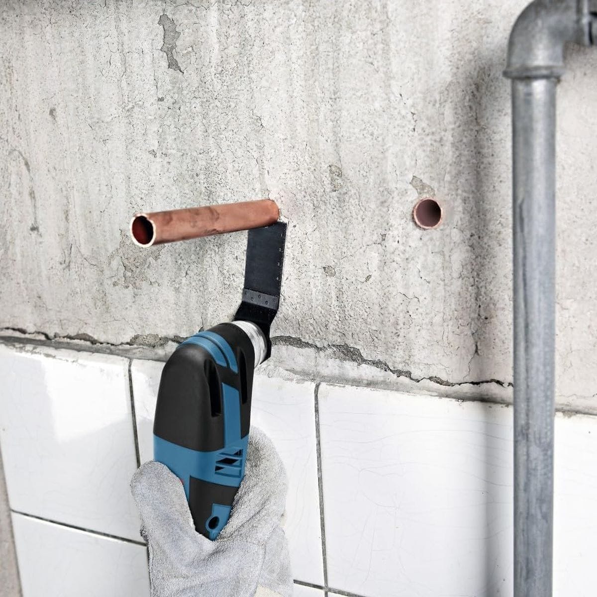 A close-up application shot of a tradesperson utilizing the Bosch cordless multi-tool with a Starlock plunge cutting saw blade to execute a precise, flush cut on a copper pipe protruding from an unfinished concrete wall above a tile line.
