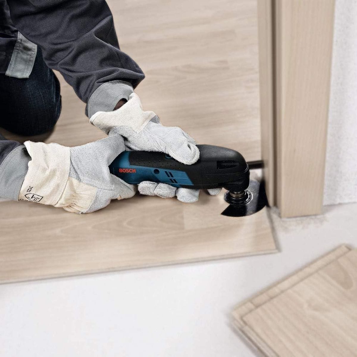 An operational action view of a flooring installer wearing protective gloves holding the compact Bosch GOP12V-28 oscillating tool flat against laminate flooring to cleanly flush-cut the base of a wooden doorjamb with a segment saw blade.