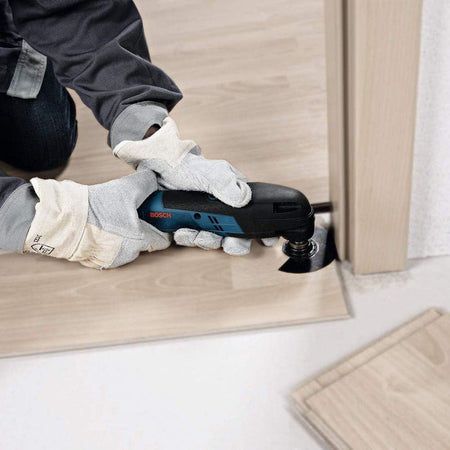 An operational action view of a flooring installer wearing protective gloves holding the compact Bosch GOP12V-28 oscillating tool flat against laminate flooring to cleanly flush-cut the base of a wooden doorjamb with a segment saw blade.