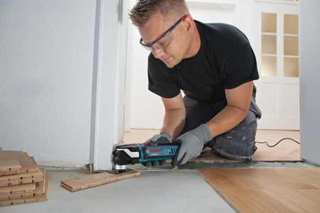 Individual using Bosch oscillating multi-tool to trim wood flooring near wall during installation project.