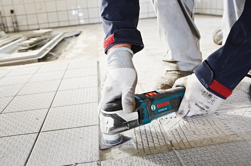 Person using Bosch multi-tool to remove tile adhesive from floor; gloves and construction materials visible.