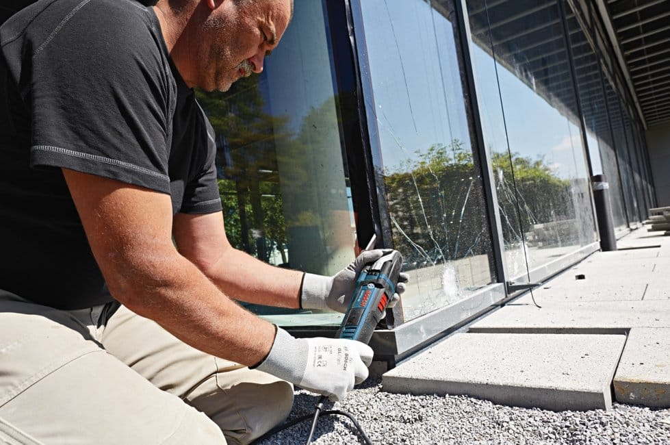 Individual kneeling outdoors using Bosch power tool near cracked glass window; gloves and gravel surface visible.