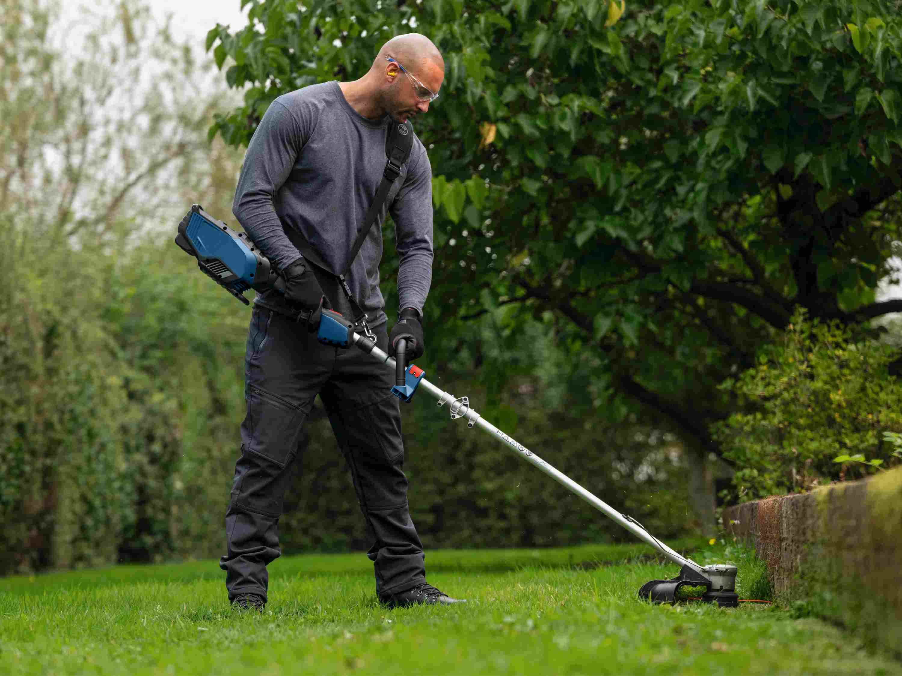 A wide lifestyle shot of a landscaper using the Bosch GRT18V-40 string trimmer with the included shoulder strap, showcasing the tool's ergonomic D-handle and balanced operation on a large lawn.
