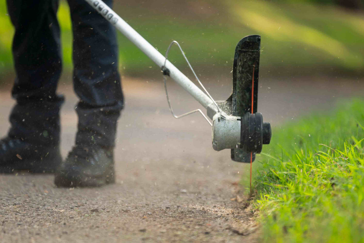 A close-up action shot of the Bosch 18V brushless string trimmer performing precision vertical edging along a concrete pathway, showcasing the high-speed 5,300 RPM cutting performance and specialized debris guard.