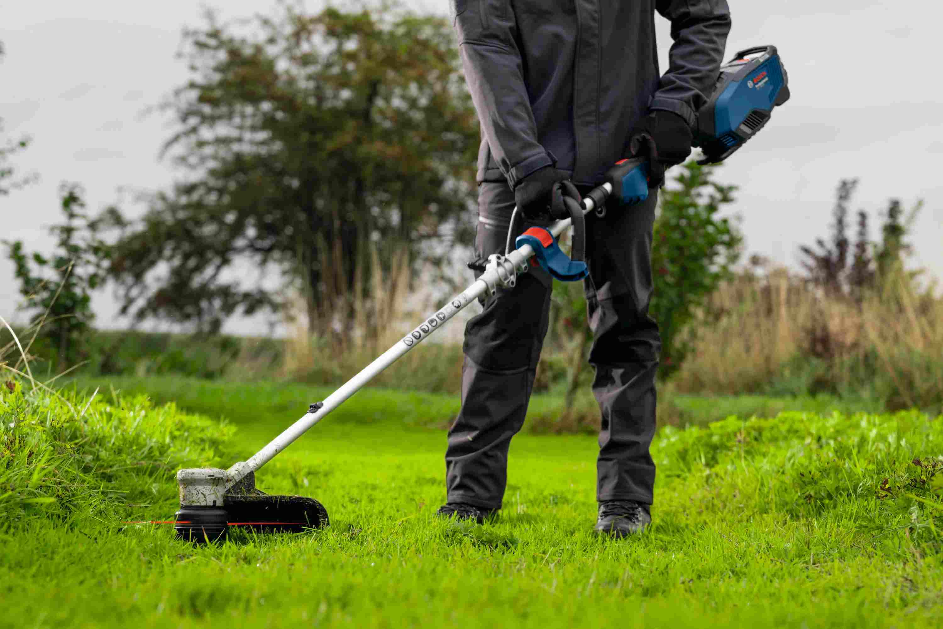 A professional landscaper using the Bosch 16 inch string trimmer to clear thick grass in an open field, highlighting the tool's balanced design and ergonomic D-handle that enhances operator comfort during long applications.