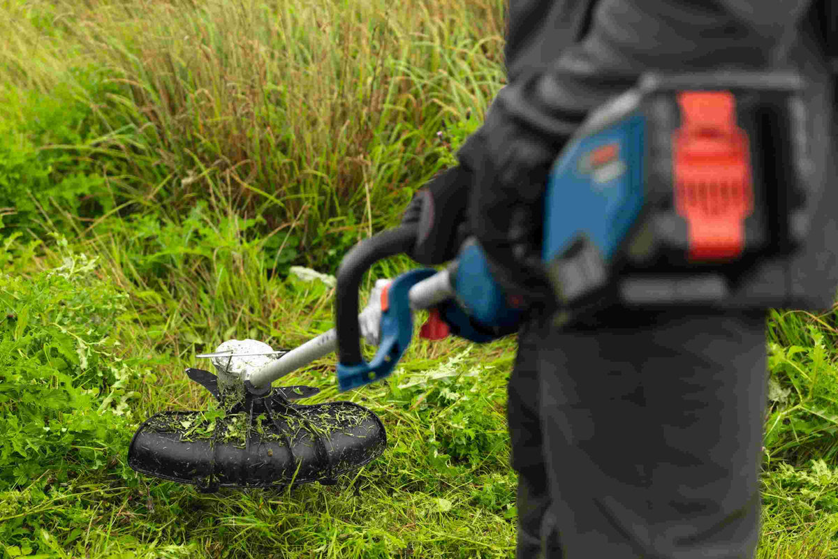 An operator using the Bosch cordless string trimmer to cut through dense, overgrown weeds, showcasing the anti-tangle technology and industrial-grade aluminum motor housing built for heavy-duty landscaping tasks.
