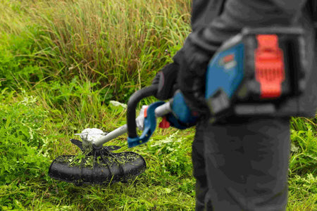 An operator using the Bosch cordless string trimmer to cut through dense, overgrown weeds, showcasing the anti-tangle technology and industrial-grade aluminum motor housing built for heavy-duty landscaping tasks.