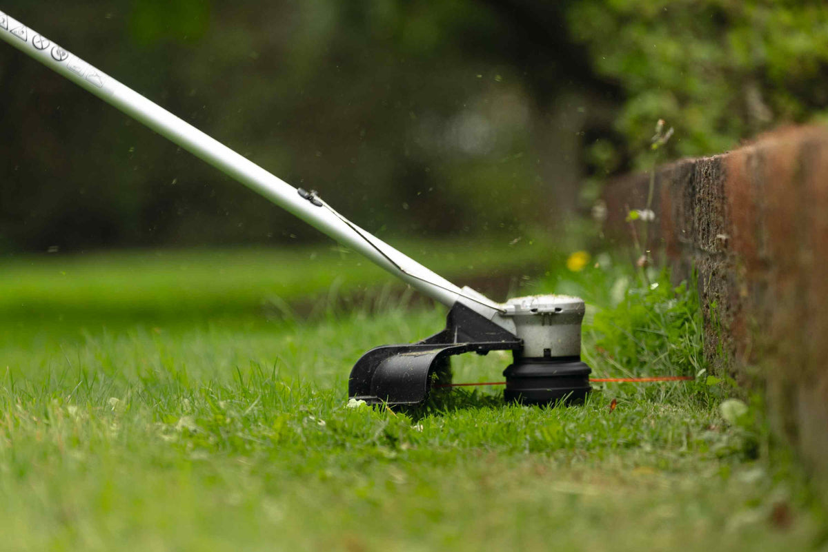 A close-up view of the Bosch trimmer head performing precise edging against a brick retaining wall, highlighting the high-speed 5,300 RPM performance and clean cutting capabilities.