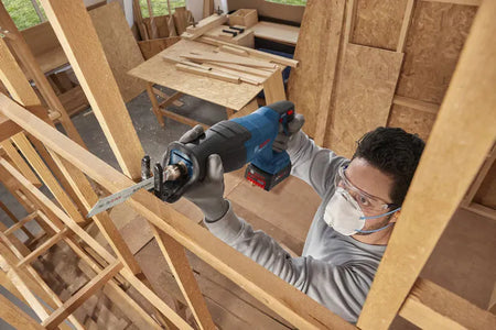 An overhead action shot of a trade professional wearing a protective respiratory dust mask and safety glasses using the Bosch GSA18V-280N cordless brushless reciprocating saw to plunge-cut through wooden stud framing. The worker guides the heavy duty demolition saw with both hands inside a timber home construction framing site.
