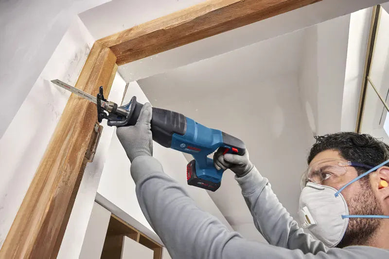 A close-up action view of a carpenter wearing a safety mask and protective eyewear utilizing the Bosch GSA18V-280N 18V max reciprocating saw to modify a wooden door frame header. The compact, handheld demolition saw is held overhead with its steel pivoting shoe stabilizing the tool against the raw timber workpiece.