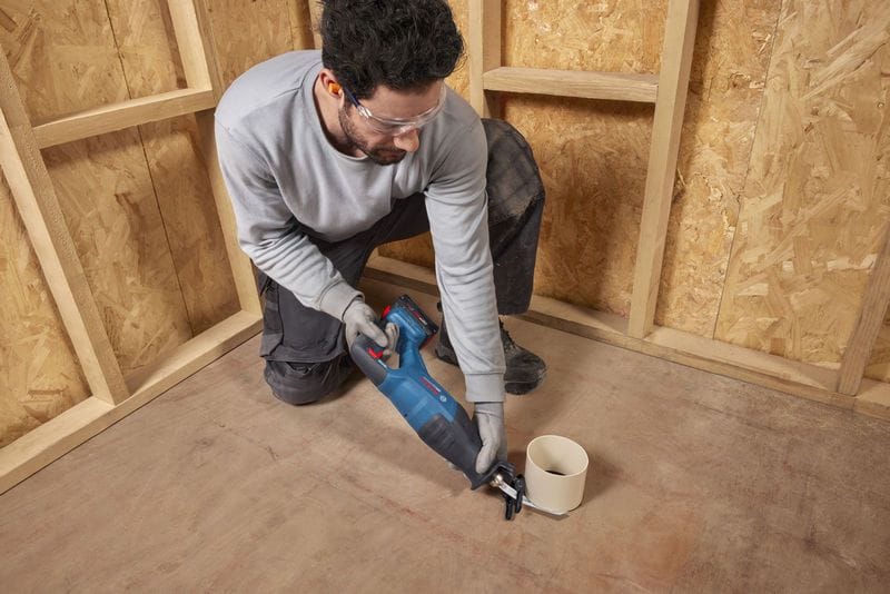A mechanical technician crouching down on a subfloor to trim a vertical white PVC plumbing drainage pipe with the Bosch GSA18V-280N brushless reciprocating saw. The worker handles the cordless cutting tool with steady manual coordination, demonstrating its low-vibration utility for tight, close-quarters pipe fitting.