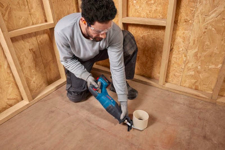 A mechanical technician crouching down on a subfloor to trim a vertical white PVC plumbing drainage pipe with the Bosch GSA18V-280N brushless reciprocating saw. The worker handles the cordless cutting tool with steady manual coordination, demonstrating its low-vibration utility for tight, close-quarters pipe fitting.