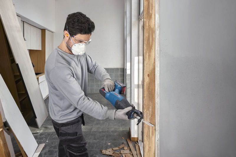 A field installation specialist equipped with a safety dust mask and clear protective glasses utilizing the Bosch GSA18V-280N cordless demolition saw to cut down a vertical wood wall framing piece. The uncropped shot showcases the balanced power-to-weight layout of the tool during a home remodeling project.
