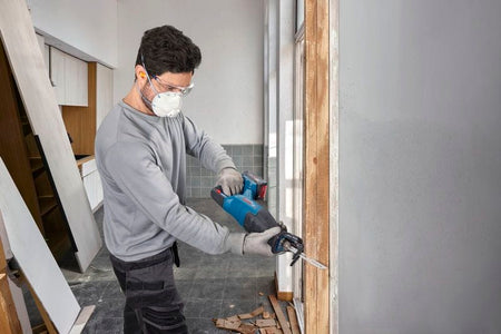A field installation specialist equipped with a safety dust mask and clear protective glasses utilizing the Bosch GSA18V-280N cordless demolition saw to cut down a vertical wood wall framing piece. The uncropped shot showcases the balanced power-to-weight layout of the tool during a home remodeling project.