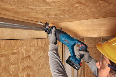 An action shot identical to file GSA18V-280N 3.jpg capturing a field technician wearing a yellow safety hard hat and clear glasses as they handle the Bosch 18V cordless reciprocating saw to cut an iron overhead pipe run. This graphic-free variant emphasizes the compact power-to-weight balance and two-handed control design of the demolition saw on active construction worksites.