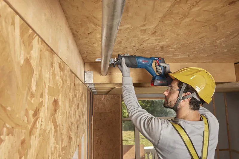 A wider workshop view identical to file GSA18V-280N 7.jpg showing a commercial plumber or electrician utilizing the Bosch GSA18V-280N brushless reciprocating saw to make a smooth slice through an overhead conduit line. The uncropped shot details the expansive wooden building structural frames and ambient daylight illuminating the craftsman's workspace.