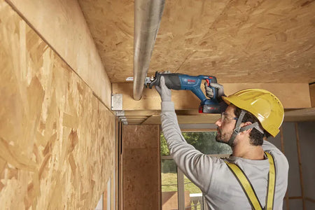 A wider workshop view identical to file GSA18V-280N 7.jpg showing a commercial plumber or electrician utilizing the Bosch GSA18V-280N brushless reciprocating saw to make a smooth slice through an overhead conduit line. The uncropped shot details the expansive wooden building structural frames and ambient daylight illuminating the craftsman's workspace.