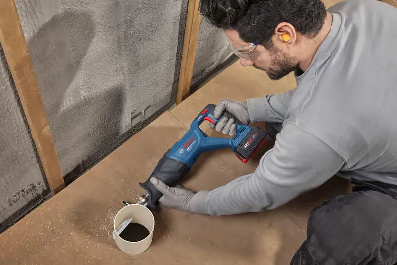A close-up view of a mechanical technician kneeling on a workshop floor to trim a white PVC plastic drainage pipe with the Bosch 18V cordless reciprocating saw bare tool. The worker guides the bi-metal demolition blade with steady manual coordination, proving the smooth low-vibration utility of the tool for close-quarters trade pipe fitting.