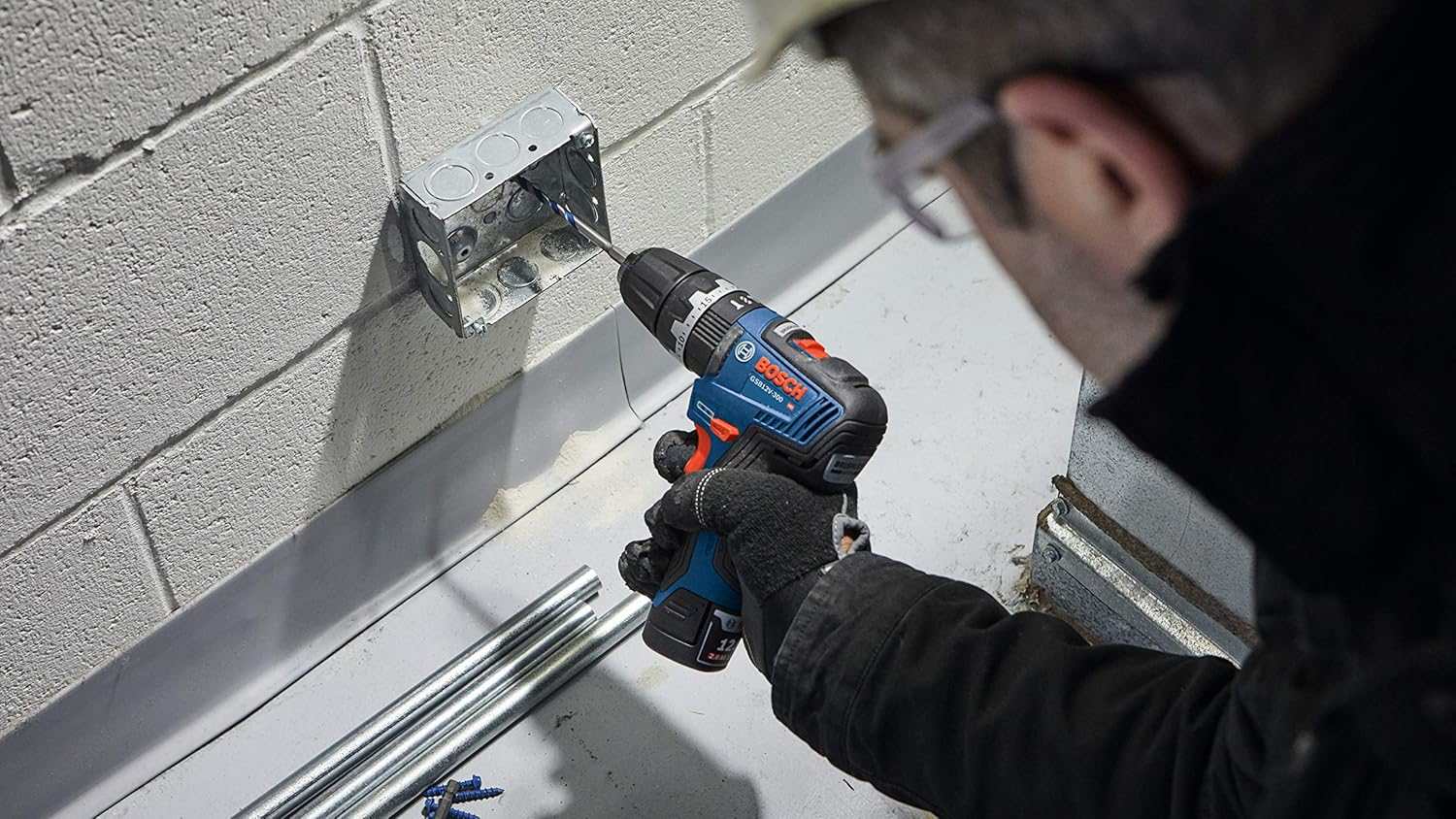 An action shot of a professional in protective gear using the compact Bosch hammer drill/driver to bore a hole into an electrical junction box mounted on a white cinder block wall.