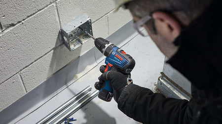 An action shot of a professional in protective gear using the compact Bosch hammer drill/driver to bore a hole into an electrical junction box mounted on a white cinder block wall.