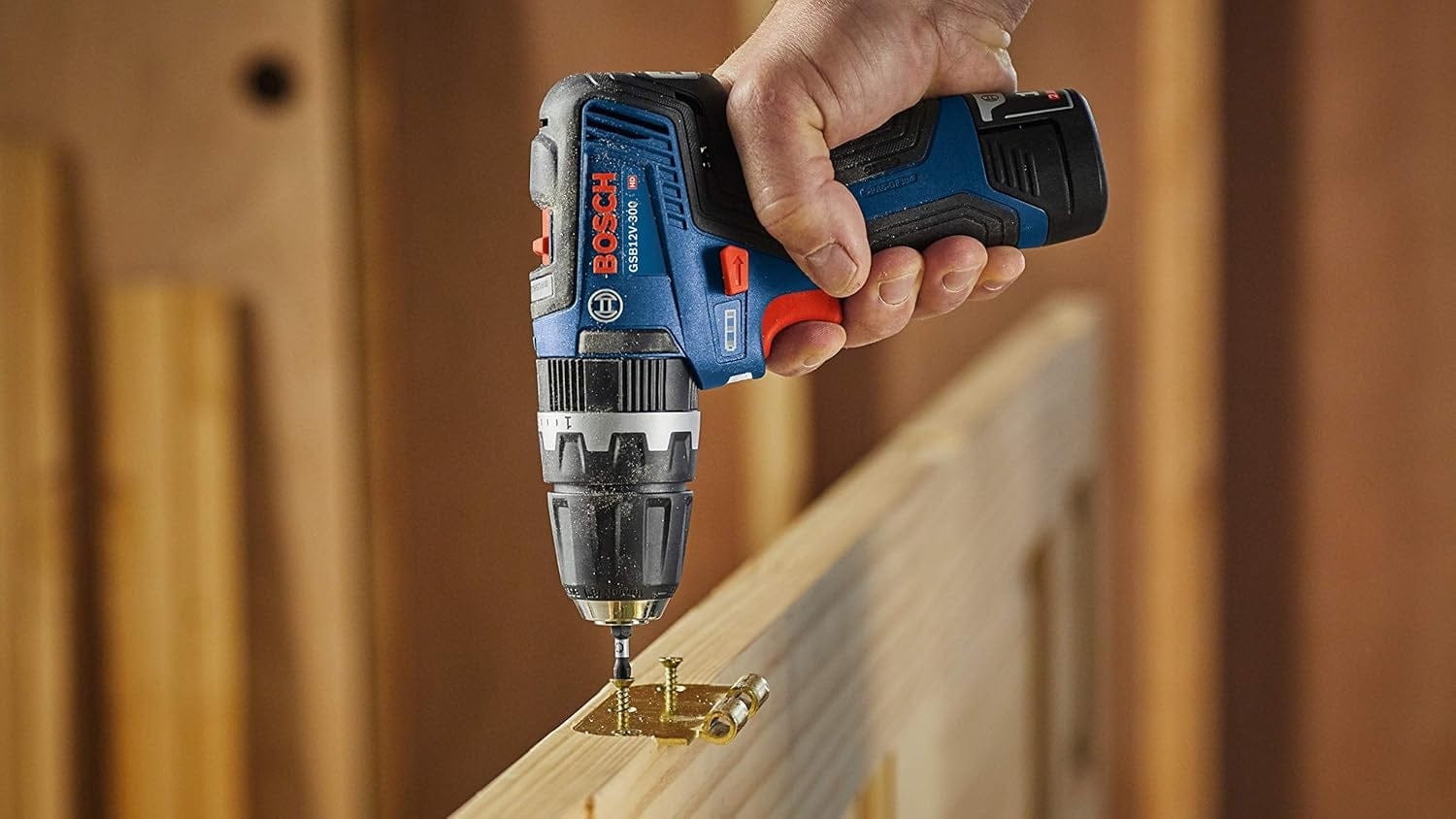 Close-up action shot showing the Bosch drill/driver being used to install a brass hinge onto the top edge of a wooden cabinet door, demonstrating its precision in delicate tasks.