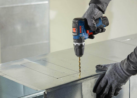 Action shot of a user wearing protective gloves using the 12V Max hammer drill/driver to bore a hole into a sheet of metal ductwork on a jobsite.