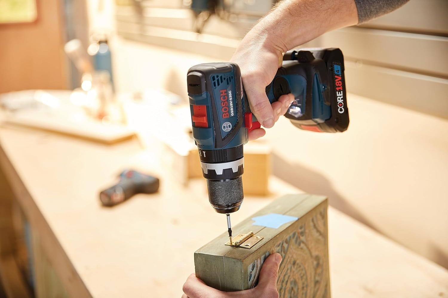 Action view of the Bosch 18V drill driver being used to fasten a hinge onto a wooden box, illustrating the tool's controlled torque for woodworking tasks.