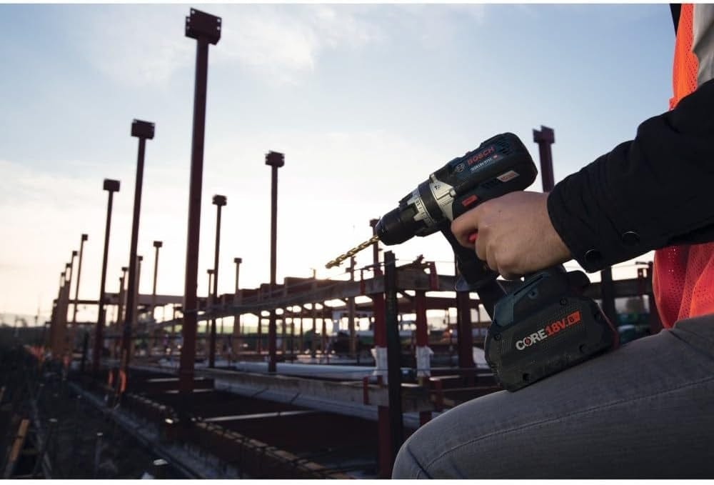 An action shot showing a professional user's hand holding the Bosch hammer drill driver with a drill bit attached, set against a construction site background with structural steel beams during sunrise or sunset.