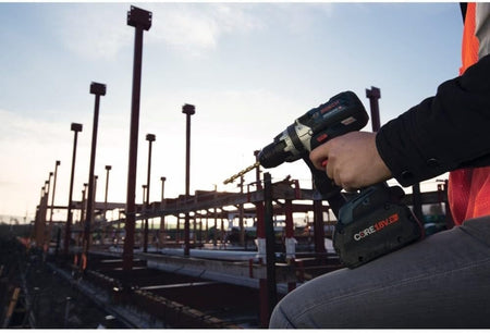 An action shot showing a professional user's hand holding the Bosch hammer drill driver with a drill bit attached, set against a construction site background with structural steel beams during sunrise or sunset.