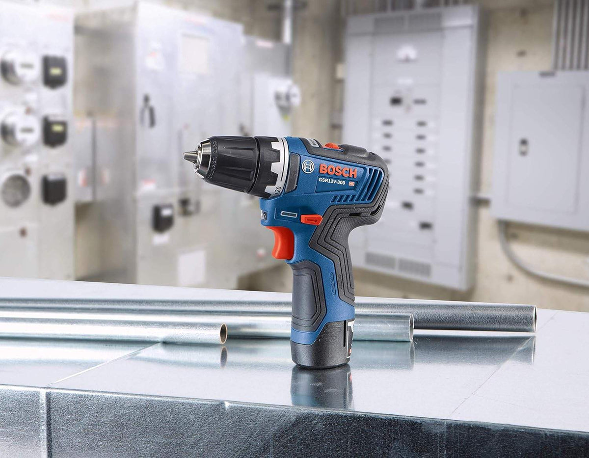 A studio shot of the Bosch GSR12V-300 drill/driver resting on a metallic surface next to sections of conduit, with an industrial electrical panel visible in the background.