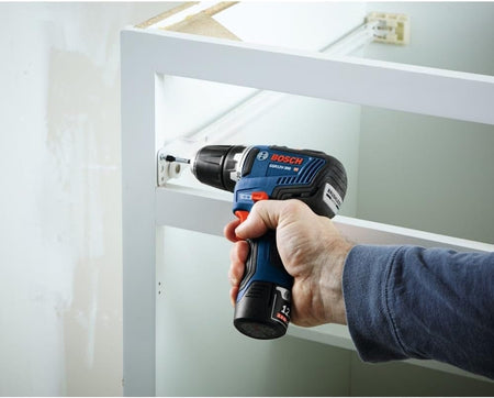 Close-up action shot showing the Bosch drill/driver being used to install hardware inside a white wooden cabinet, demonstrating its effectiveness in confined spaces.