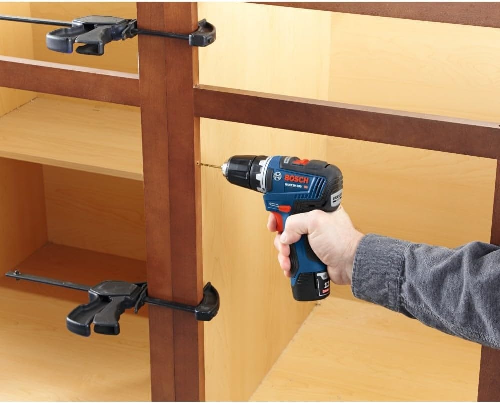 A person using the 12V Max drill to bore a pilot hole into a wooden cabinet frame that is held together by bar clamps.