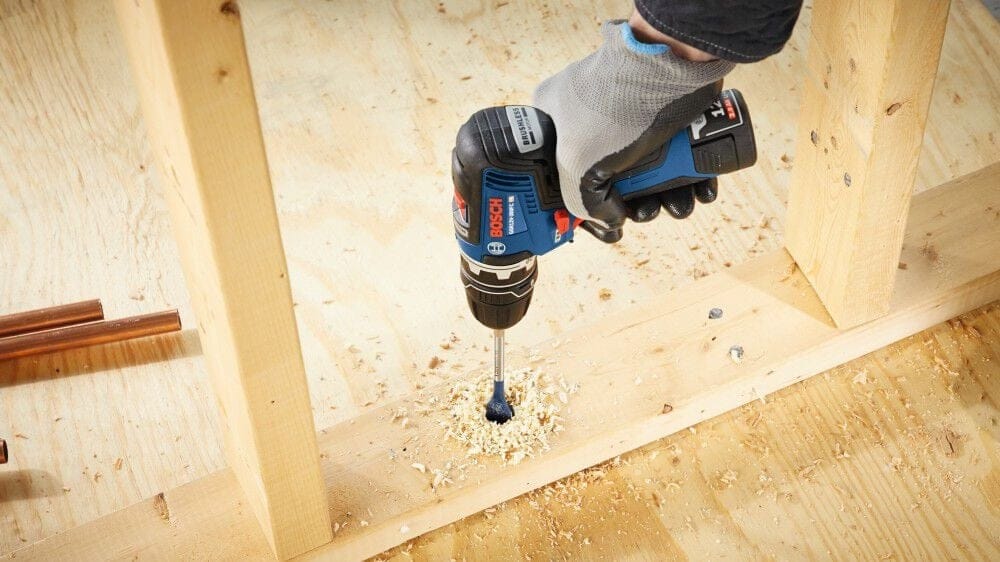 A close-up of a person wearing gloves using the Bosch 12V Max Chameleon FlexiClick 5-in-1 drill driver with the right angle attachment to drill a hole into a wooden stud near the floor.