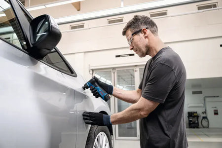 An automotive refinishing technician wearing protective clear safety glasses and black work gloves uses the Bosch GSS12V-32N 12V max cordless detail orbital sander bare tool to fix a small paint defect. The worker guides the compact brushless 1-1/4 inch sander smoothly along the side panel of a silver car inside a clean detailing garage workshop.