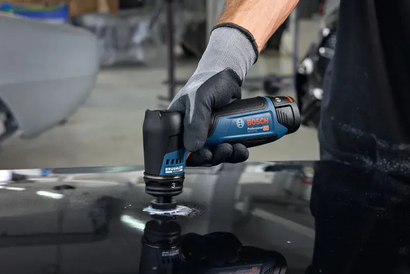 An automotive refinishing technician clad in gray and black protective gloves using the Bosch GSS12V-32N brushless detail sander to flat-level an imperfection on a mirror-like black vehicle surface. The close-up action view shows the micro 1-1/4 inch pad working directly through a dollop of white dry-sanding paste.