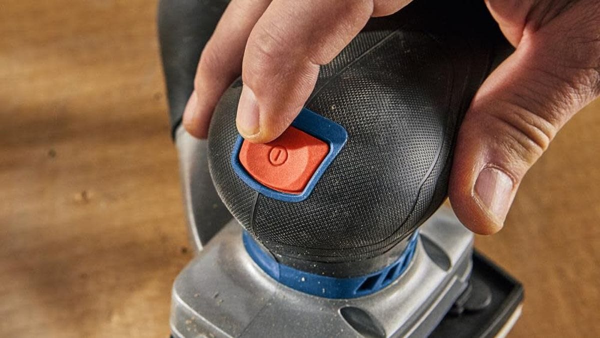 An overhead action view of a woodworker pressing down on the textured palm rest of the Bosch GSS18V-40 orbital sander, using the triangular detail sanding plate configuration to smooth an internal corner of a hardwood staircase step.