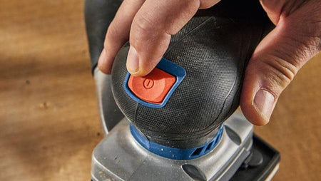 An overhead action view of a woodworker pressing down on the textured palm rest of the Bosch GSS18V-40 orbital sander, using the triangular detail sanding plate configuration to smooth an internal corner of a hardwood staircase step.