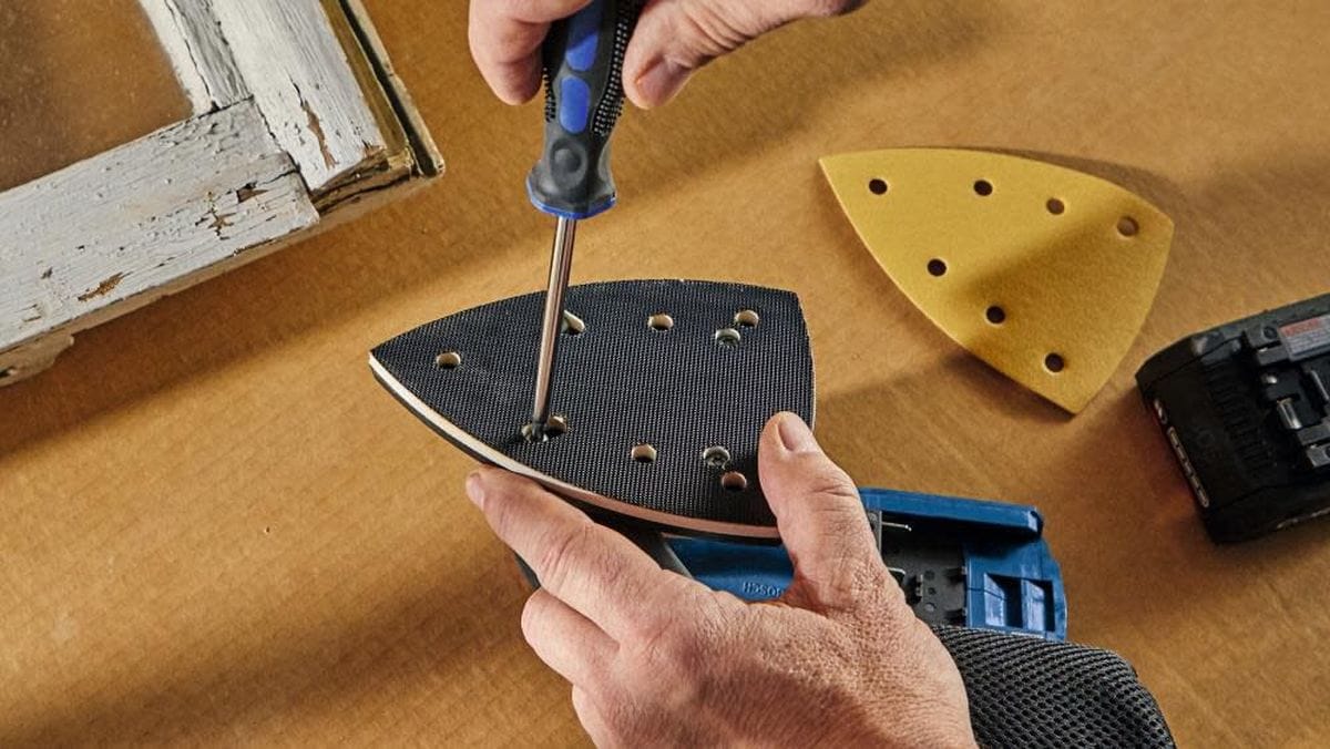 A close-up view of a technician using a hand screwdriver to change out the interchangeable base on the Bosch cordless sander, fastening a pointed detail sanding plate into position on a workbench next to a yellow sheet of multi-hole abrasive sandpaper.