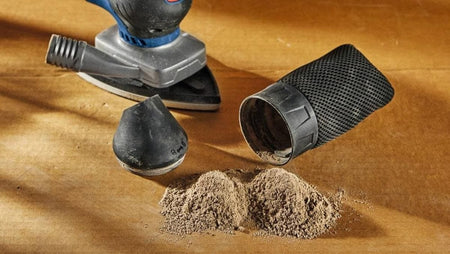A product component shot showing a small pile of collected fine wood dust on a work table next to the detached fabric dust extraction collection bag and universal vacuum hose adapter accessory from the Bosch sander kit.