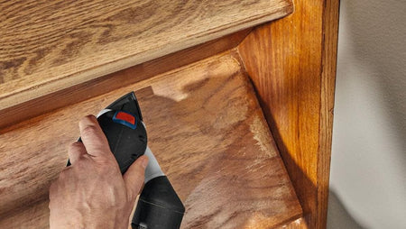 An overhead perspective view of a trade professional guiding the compact cordless Bosch palm sander along the grain of a vertical wooden staircase panel, utilizing the detail sheet configuration to strip old varnish.