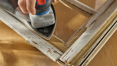 An active jobsite view showing a craftsman using the Bosch 18V brushless finishing sander to carefully remove peeling white paint and prep the detailed wooden trim molding around a vintage glass window frame.